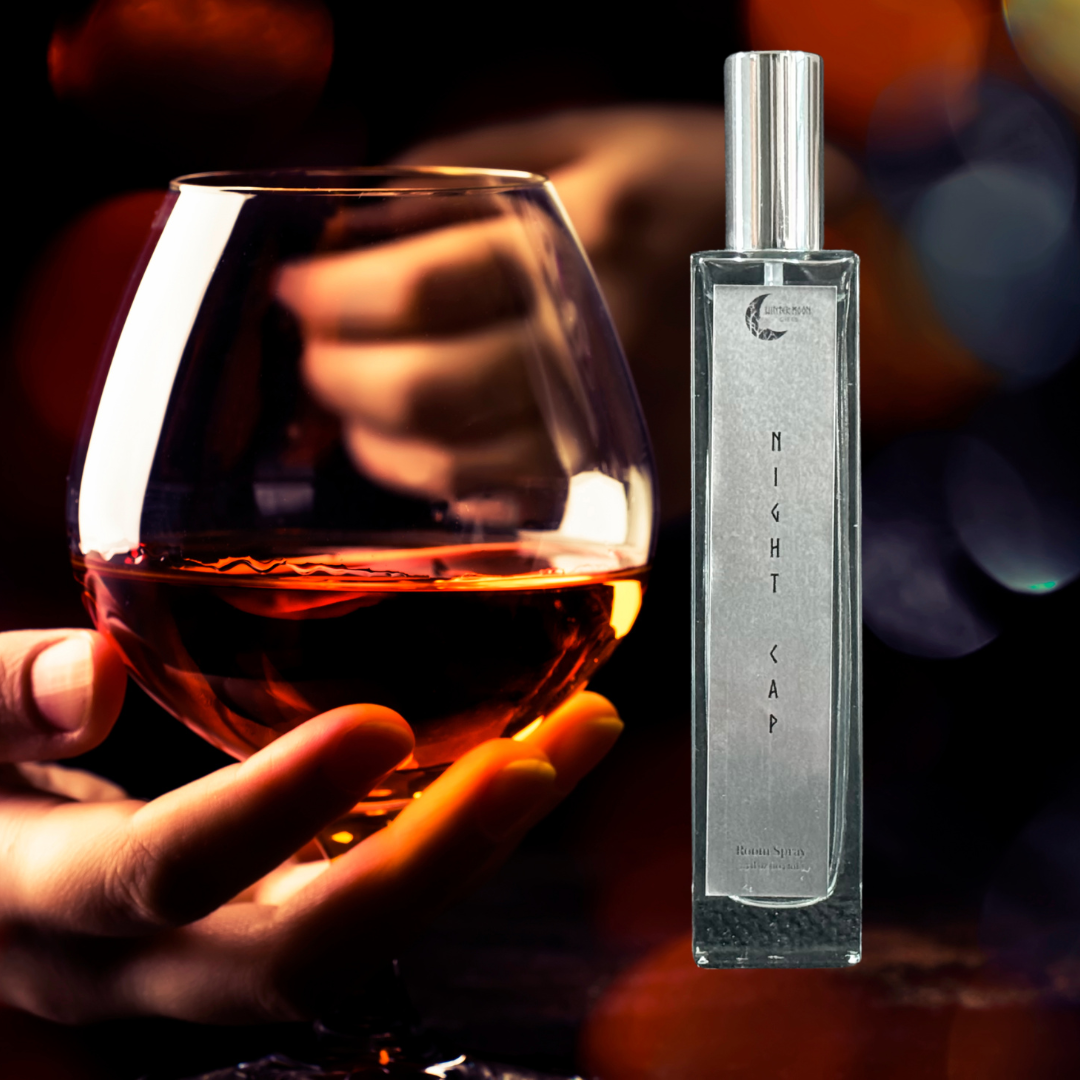 Night Cap Room Spray bottle in a dim, moody setting with a hand holding a glass of cognac and warm bokeh lighting in the background.